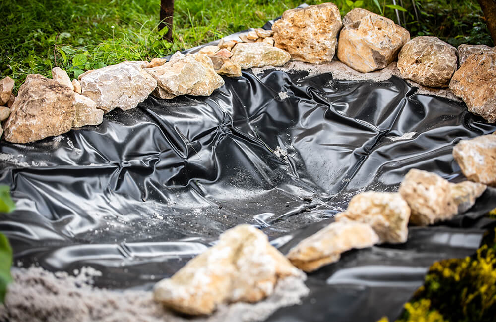 A pond is being prepared with pond liner and some stones to fixate it.